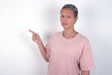 Serious young woman with short hair wearing pink t-shirt over white background smirks face points away on copy space shows something unpleasant. Look at this advertisement. Big price concept