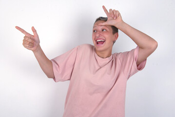 young woman with short hair wearing pink t-shirt over white background showing loser sign and...