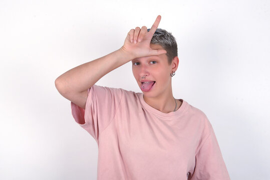 Funny young woman with short hair wearing pink t-shirt over white background makes loser gesture mocking at someone sticks out tongue making grimace face.