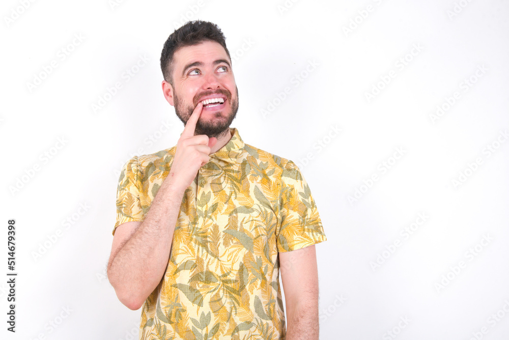 Wall mural Young caucasian man wearing Hawaiian t-shirt over white background  with thoughtful expression, looks to the camera, keeps hand near face, bitting a finger thinks about something pleasant.