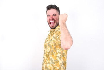 Overjoyed Young caucasian man wearing Hawaiian t-shirt over white background  glad to receive good news, clenching fist and making winning gesture.
