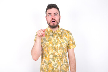 Fototapeta premium Shocked Young caucasian man wearing Hawaiian t-shirt over white background points front with index finger at camera and. Surprise and advertisement concept.
