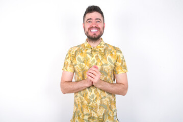 Happy young handsome bearded man wearing flowered shirt over white wall stands against orange studio wall keeps hands on heart, swears be loyal, expresses gratitude. Honesty concept.