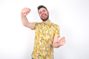 Portrait of charming young handsome bearded man wearing flowered shirt over white wall, smiling broadly while holding hands over her head.  Confidence and relax concept.