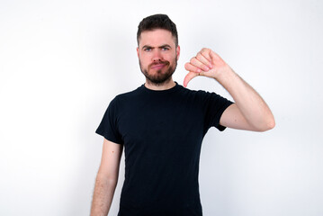 young caucasian bearded man wearing black t-shirt standing over white wall looking unhappy and angry showing rejection and negative with thumbs down gesture. Bad expression.