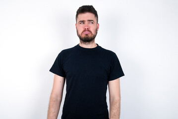 Dissatisfied young caucasian bearded man wearing black t-shirt standing over white wall purses lips and has unhappy expression looks away stands offended. Depressed frustrated model.