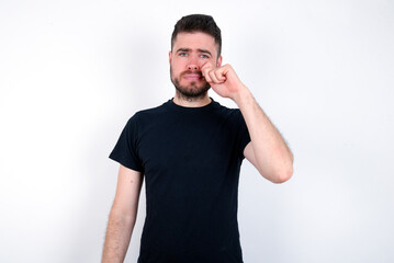 Disappointed dejected young caucasian bearded man wearing black t-shirt standing over white wall wipes tears stands stressed with gloomy expression. Negative emotion