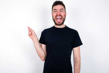 young caucasian bearded man wearing black t-shirt standing over white wall points aside on copy blank space. People promotion and advertising concept