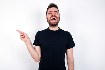 young caucasian bearded man wearing black t-shirt standing over white wall laughs happily points away on blank space demonstrates shopping discount offer, excited by good news or unexpected sale.