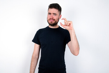 Displeased young caucasian bearded man wearing black t-shirt standing over white wall shapes little hand sign demonstrates something not very big. Body language concept.