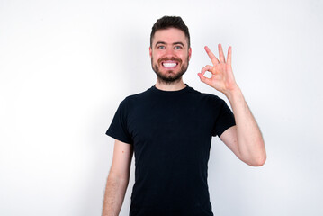young caucasian bearded man wearing black t-shirt standing over white wall hold hand arm okey symbol toothy approve advising novelty news