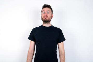 Obraz premium Stunned young caucasian bearded man wearing black t-shirt standing over white wall stares reacts on shocking news. Astonished man holds breath