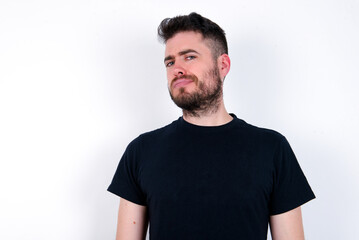 Fototapeta premium Displeased young caucasian bearded man wearing black t-shirt standing over white wall frowns face feels unhappy has some problems. Negative emotions and feelings concept