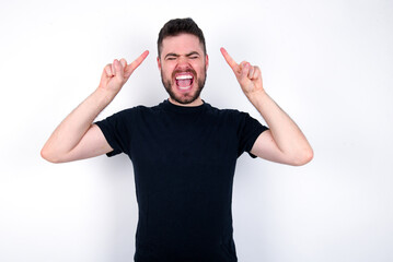 Fototapeta premium Photo of crazy young caucasian bearded man wearing black t-shirt standing over white wall screaming and pointing with fingers at hair closed eyes