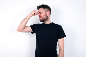 Fototapeta premium Sad young caucasian bearded man wearing black t-shirt standing over white wall suffering from headache holding hand on her face