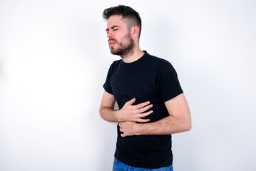 young caucasian bearded man wearing black t-shirt standing over white wall got stomachache