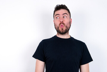 Shocked young caucasian bearded man wearing black t-shirt standing over white wall look empty space with open mouth screaming: Oh My God! I can't believe this.