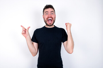 Fototapeta premium young caucasian bearded man wearing black t-shirt standing over white wall points at empty space holding fist up, winner gesture.