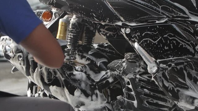 Worker doing detailed cleaning and washing a motorbike with foam soap in car wash station.