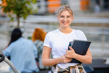 Portrait of a Beautiful smiling blonde hair student girl with glasses  holding books in front of a university building - Powered by Adobe