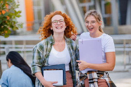 Couple of cheerful female university students in front off university building holding books, smiling