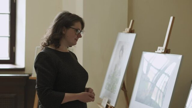 Young thoughtful woman in glasses with curly hair visits art gallery. Lady in black plaid dress, with smile and pleasure examines paintings on easels at exhibition. Reflection from window.