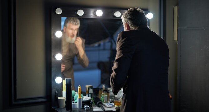 Handsome gray haired middle aged man wearing elegant suit looking at himself in the mirror while preparing for studio photoshoot