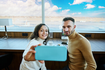 Attractive woman sitting next to her husband and taking a selfie against panoramic windows overlooking the runway while waiting to board a flight