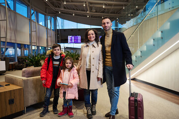 Happy multi-ethnic family with suitcases, standing against flight information board with timetable in international airport departures terminal