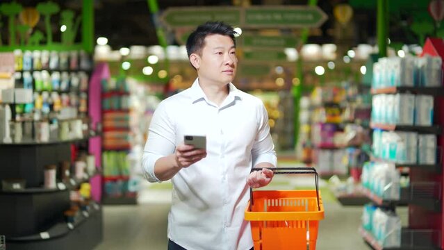asian male shopper walks around the supermarket with a shopping cart looking at smart phone browsing smartphone in hands search goods or products on the list. customer purchaser man in a grocery store