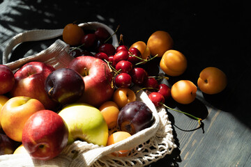 Fresh ripe fruits and berries on a black wooden background in an ecological mesh bag. Lifestyle. Hard shadows.Sunlight.Ripe harvest of apples. cherries, apricots and plums. Copy space.