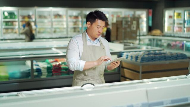 Asian grocery store manager salesman in apron using digital tablet counting goods in supermarket. Male worker employee doing inspection of products regular inventory to control goods, online order