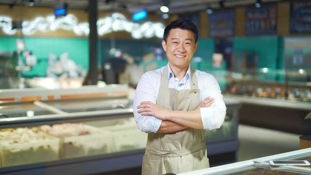 portrait of handsome asian man salesman in apron standing and looking at camera in grocery store supermarket. Male worker seller or small business owner. indoor. food, groceries market Cheerful smile