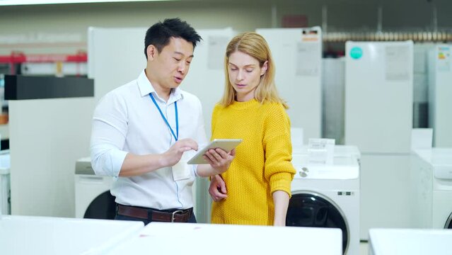 Asian male consultant Salesman Gives Professional advises female home appliance buyer in store. Shop assistant explaining washing machine to customer. showing modern electric 
