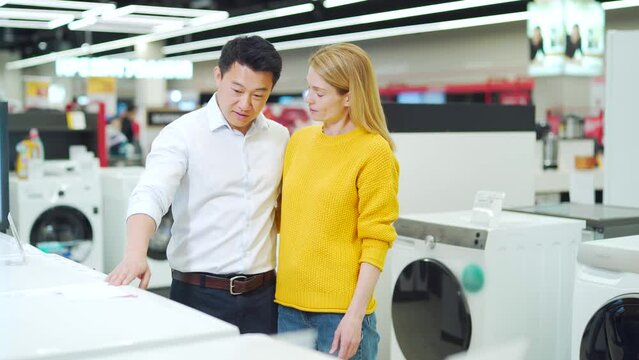 happy mixed race asian couple family man and woman make a choice choose of new household home appliances in supermarket survey cooler, freezer or washing machine store purchase and shopping