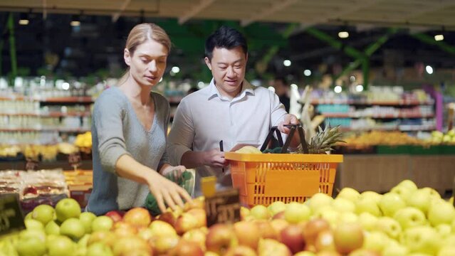 happy mixed race asian couple family man and woman choosing fruits or vegetables in grocery store supermarket. Joint daily shopping together. Buyers customer select product pick an apple in eco bag