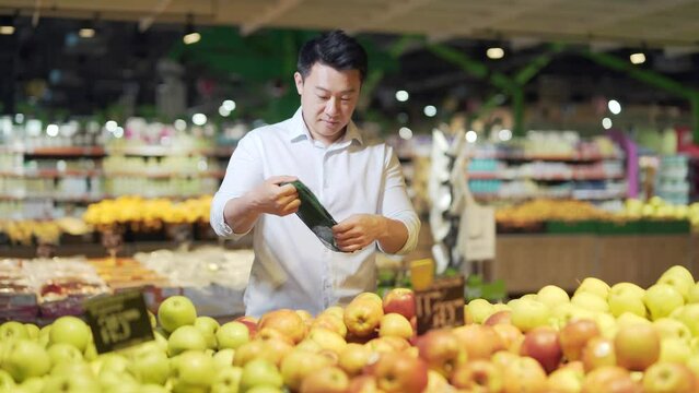 young asian man chooses and picks in eco bag apple fruit or vegetables in the supermarket. male customer standing a grocery store near the counter buys and throws in a reusable package in market