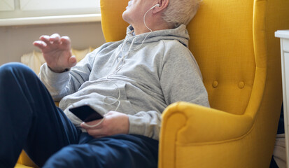 old man listens to music from a smartphone on headphones, generation, fun in solitude natural house - obrazy, fototapety, plakaty