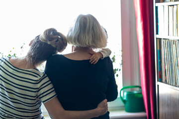 generation, family looks out the window, daughter hugs mother who is holding her granddaughter, natural background at home, view from behind