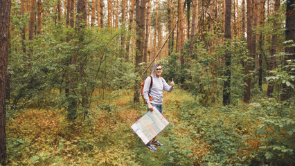 Young man in glasses with backpack and map showing gesture of like in summer forest. Male tourist showing thumb up, looking at camera, standing in woods.