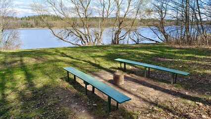 Obraz premium On the grassy bank of the river there are trees, wooden benches and a deck. On the opposite bank grows a forest. There are ripples on the water. It is sunny and the sky is blue with clouds