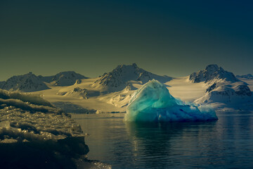 Fototapeta na wymiar 2022-07-01 A BLOCK OF FLOATING ICE IN THE ARCTIC SEA WITH A JAGGED MOUNTAIN RANGE IN THE BACKGROUND AND A PLAIN BLUE SKY