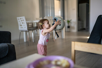 Front view portrait of one girl small caucasian toddler baby putting on wear protective helmet on head at home in room growing up learning new skills early development concept