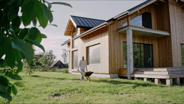Man is driving a wheelbarrow working at the backyard of a modern house on a sunny day. Man wearing a shirt and a cap. Scandinavian lifestyle man working at the backyard. High quality 4k footage