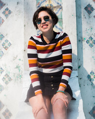 Young cheerful millennial woman posing with a striped long sleeve shirt in autumn colors, black skirt and sunglasses with a withe wall and trees in the background