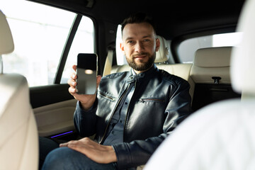 successful young man sits in the back seat of a car and demonstrates the screen of a smartphone