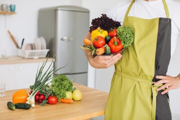 Fragment of a man holding a plate of fresh vegetables on the background of a kitchen and a kitchen table with various vegetables.