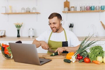 Fototapeta na wymiar Happy man in headphones preparing salad in kitchen from fresh vegetables and looking at laptop.