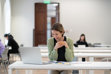 Happy female mature student sitting at library desk attending virtual class, looking at computer screen and smiling, middle-aged woman watching webinar on laptop, talking to teacher via video call