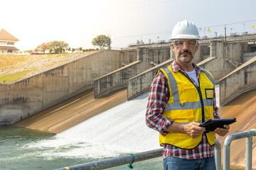 A dam engineering doing his checking routine. He is wearing a white hard hat and yellow transparent vest. He is standing by the rail by the dam.
